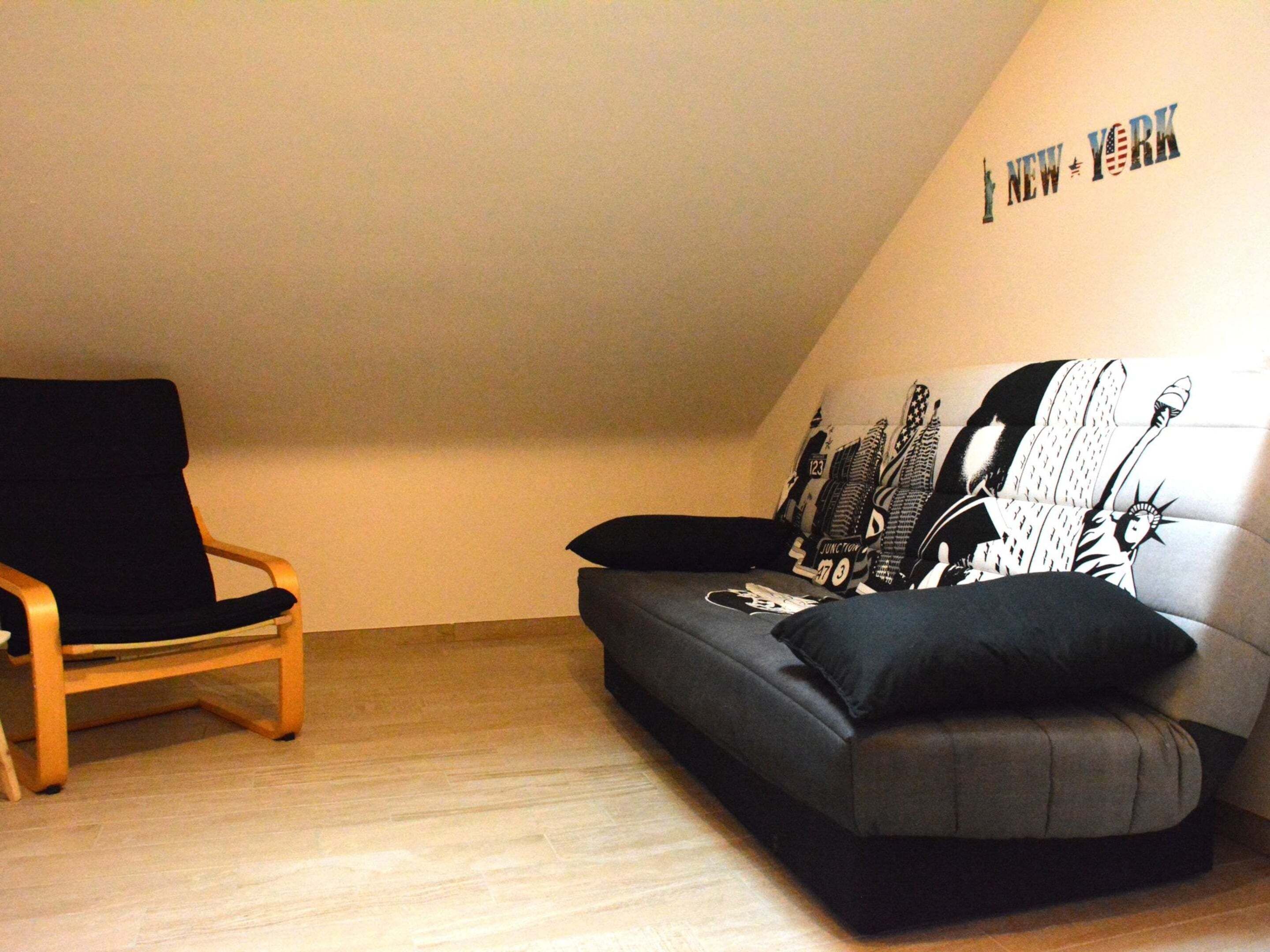 Una habitación con una silla negra, un sofá gris y una pared con 'NEW YORK' y una silueta de la Estatua de la Libertad.