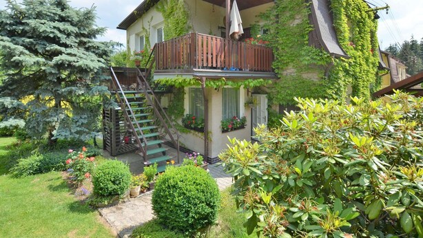 A two-story house with a wooden balcony, a staircase leading to the upper floor, and a garden with various plants and flowers. - Modern Apartment in Carlsbad With Garden (Karlovy Vary)