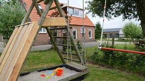 Ein Spielplatz mit einer Holzrutsche und einer Kletterkonstruktion, umgeben von Gras und einem Backsteinhaus im Hintergrund.