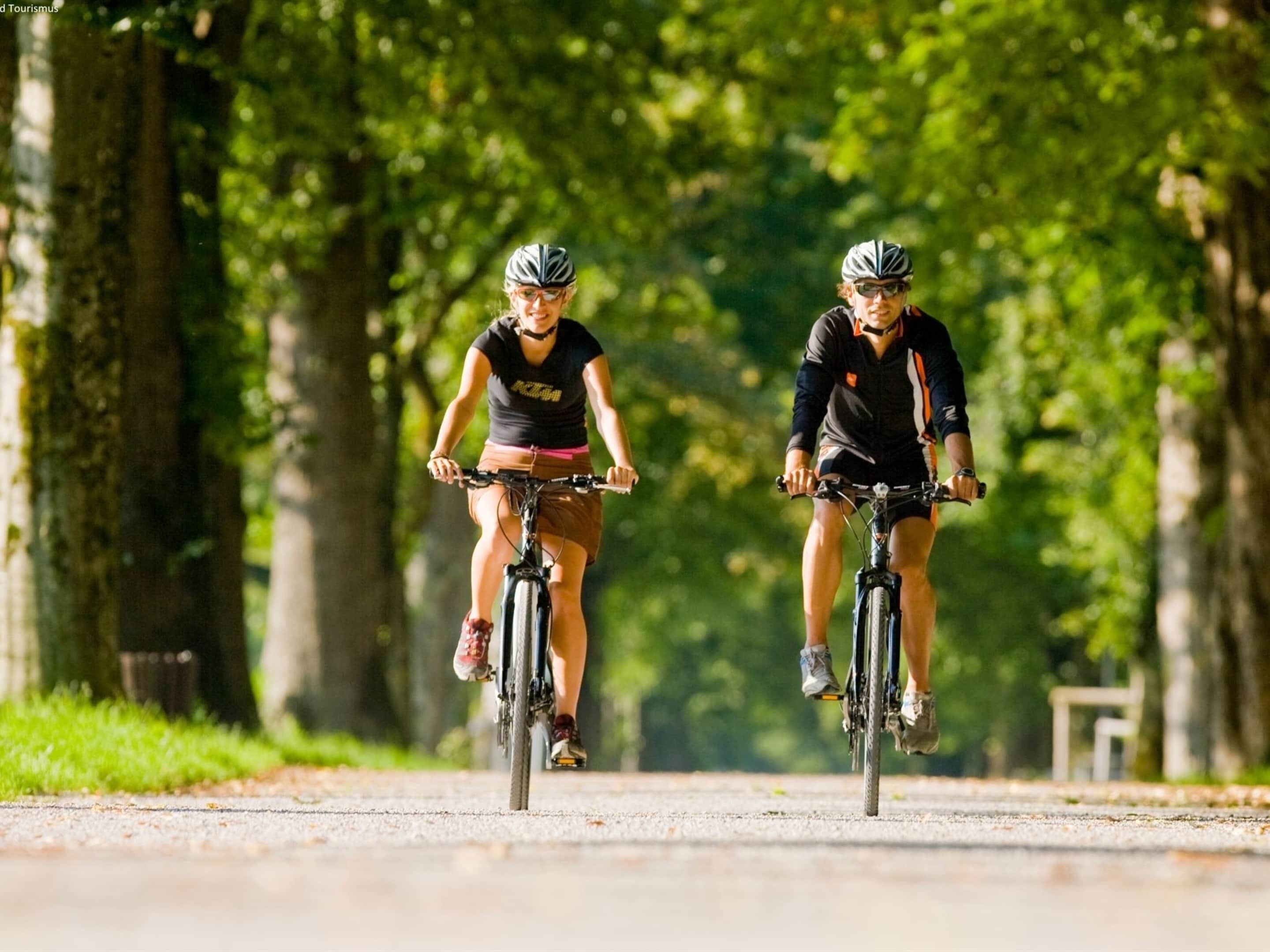 Two cyclists on a paved path surrounded by trees.
