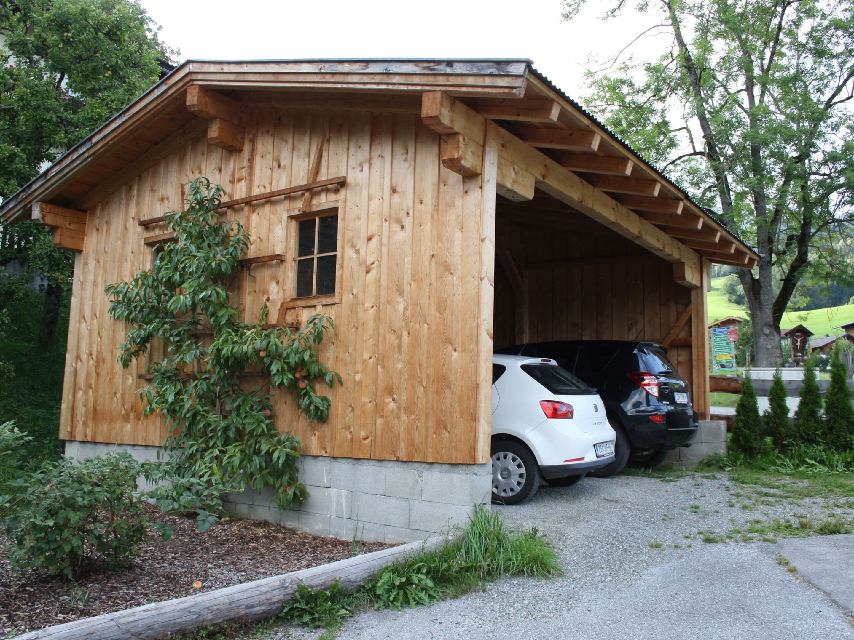 A wooden cabin with a covered entrance, two cars parked inside, and a small garden area with a wooden fence and greenery.