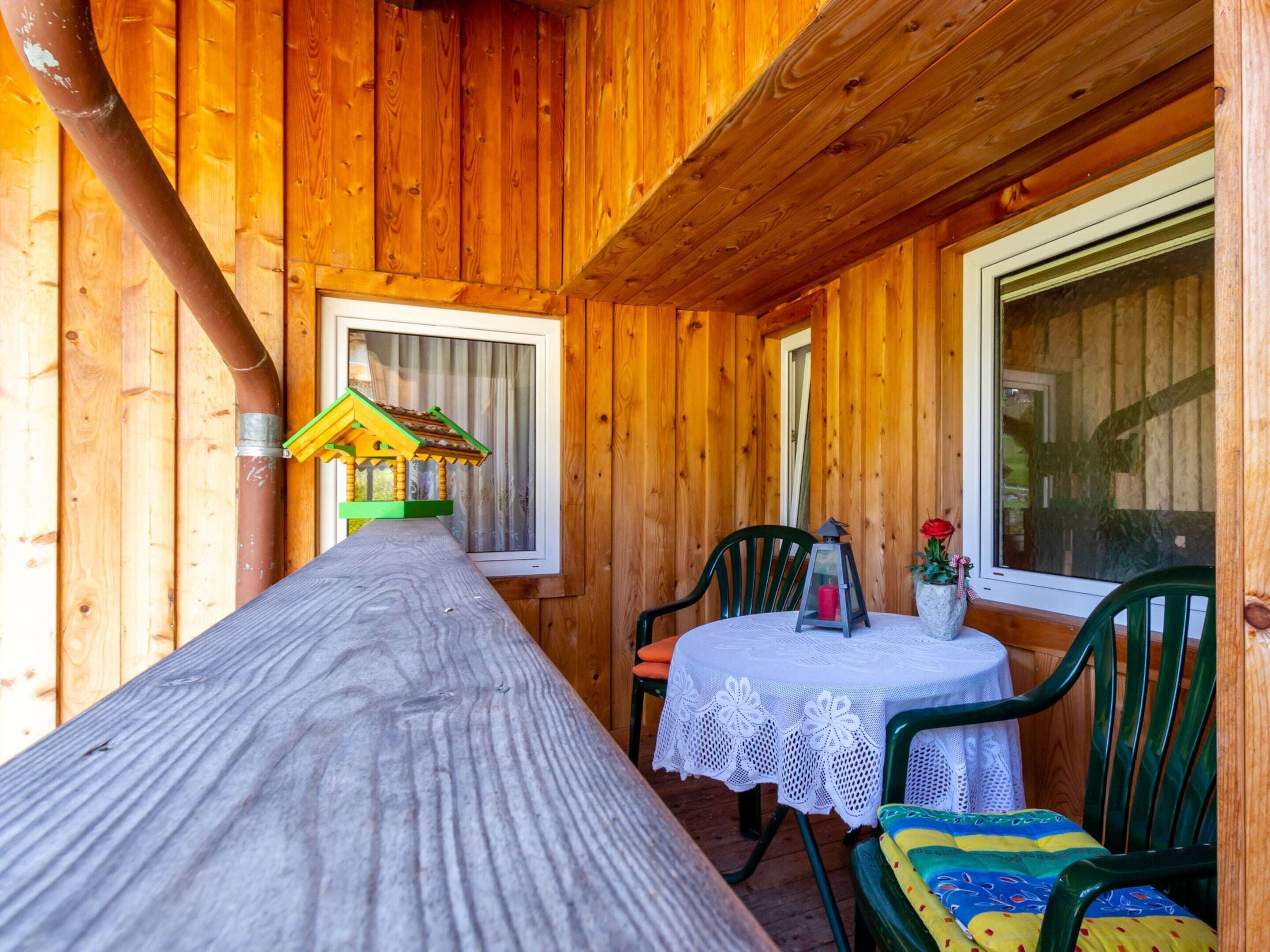 A wooden cabin interior with a long table, two chairs, a small table with a vase and a candle, and a window with curtains.