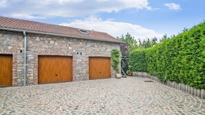 Un bâtiment en pierre avec deux portes de garage en bois, une cour pavée et une haie verte.