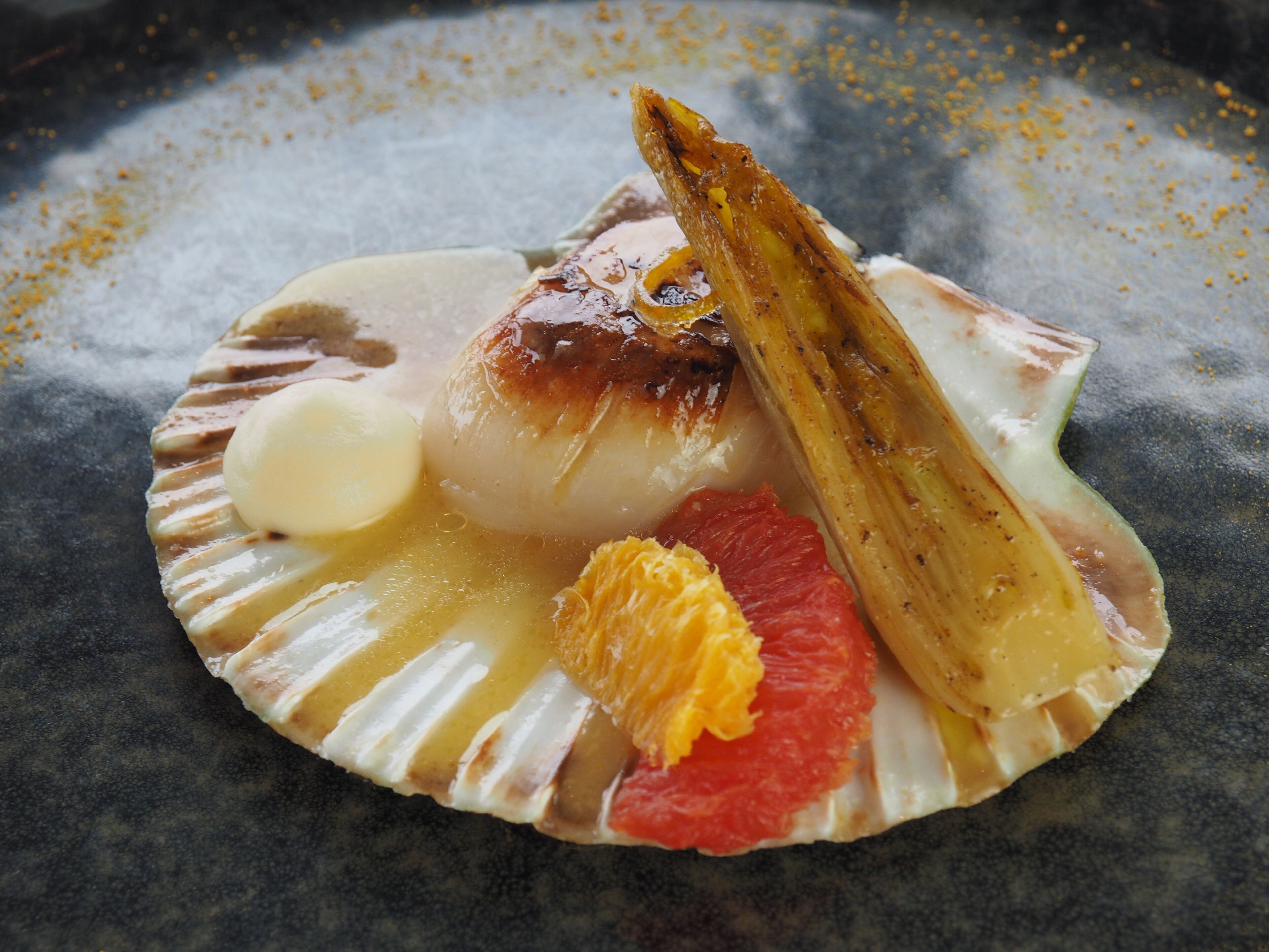 Un dessert présenté dans une assiette, avec une coquille de Saint-Jacques comme base, un élément rond de couleur blanche, un morceau de fruit et une tranche de fruit caramélisé.
