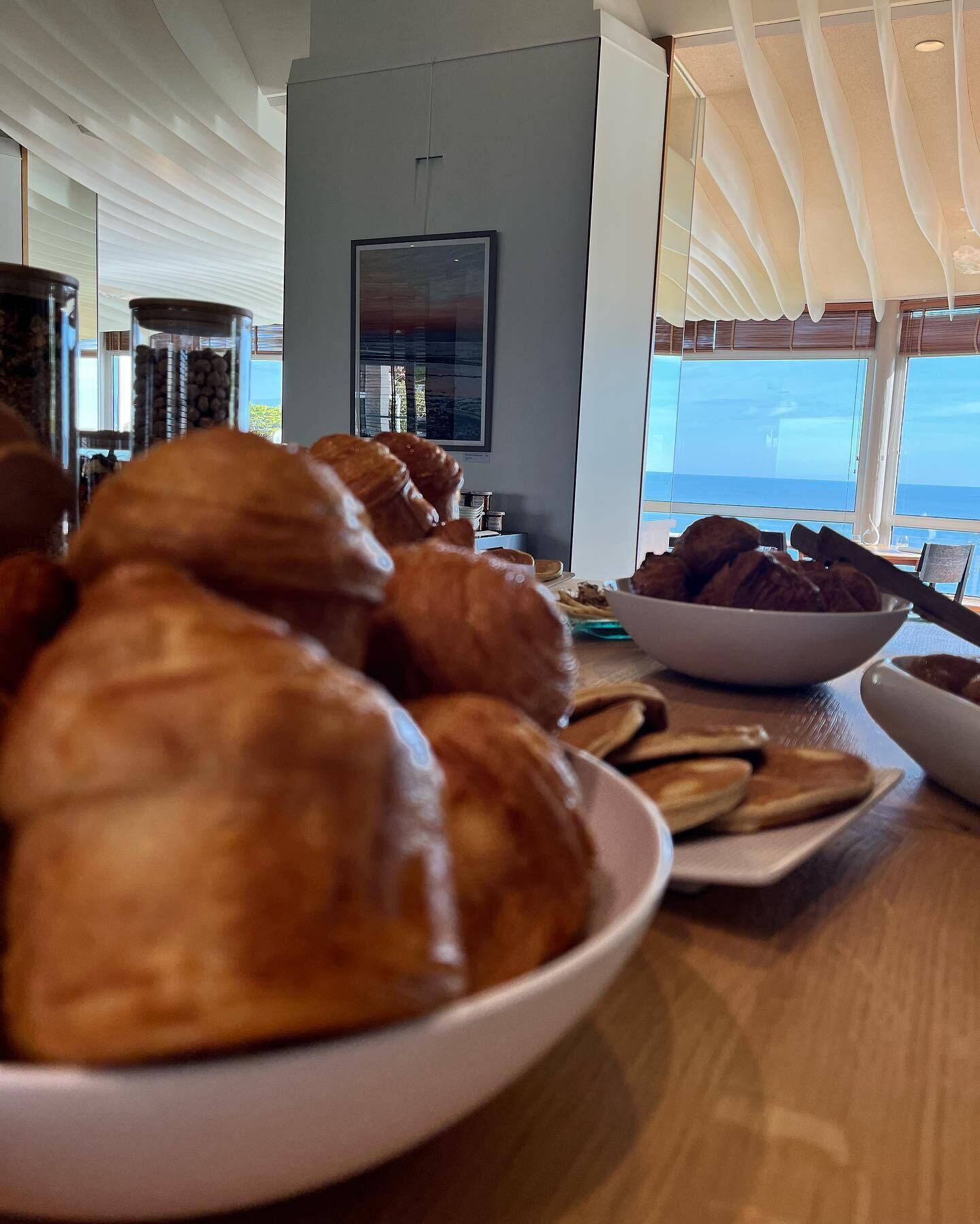 A breakfast buffet with croissants and other pastries on a wooden table, with a view of the ocean through large windows.