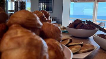 A breakfast buffet with croissants and other pastries on a wooden table, with a view of the ocean through large windows.