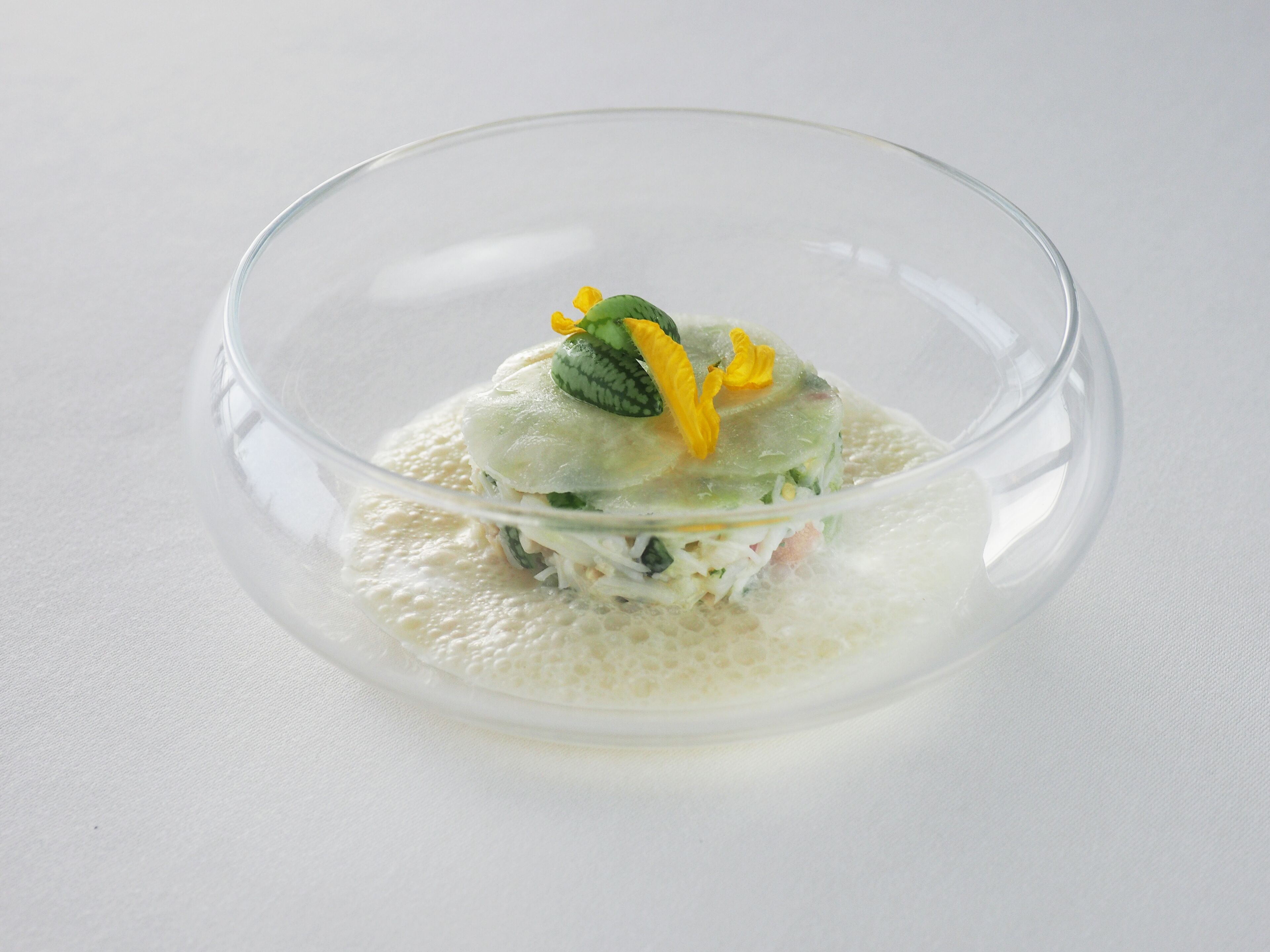 A clear glass bowl containing a layered dish with a garnish of yellow flowers and green leaves.