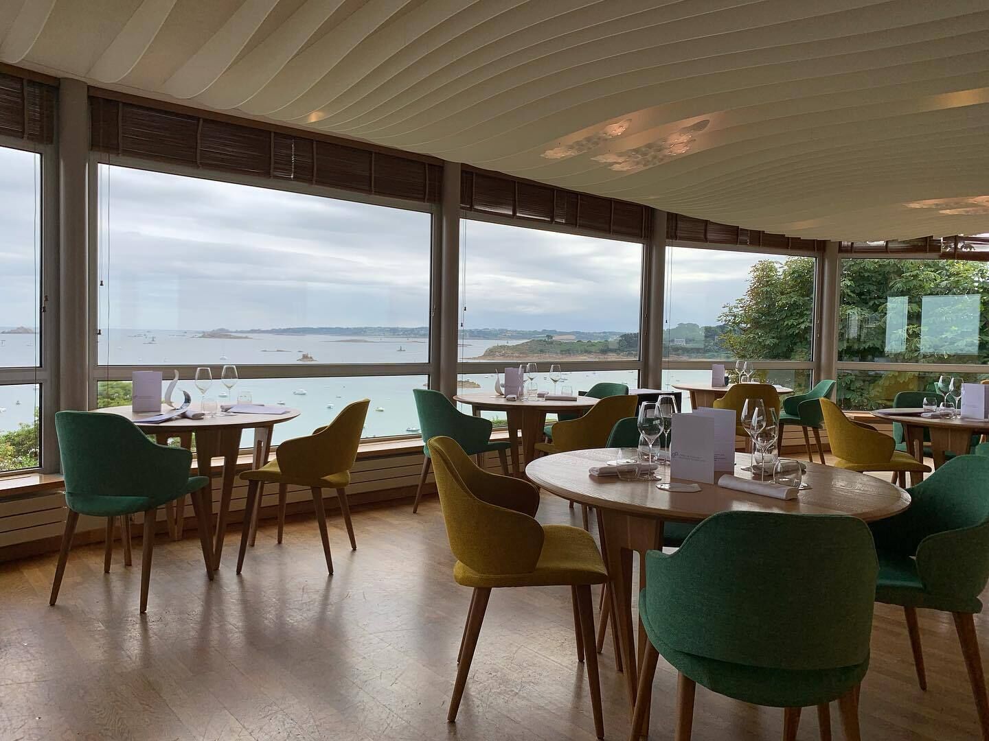 Un restaurante con mesas y sillas redondas, grandes ventanales con vistas al mar y barcos, y un piso de madera.