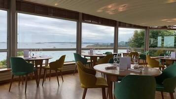 Un restaurante con mesas y sillas redondas, grandes ventanales con vistas al mar y barcos, y un piso de madera.