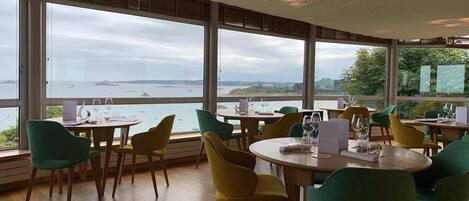 A restaurant with round tables and chairs, large windows with a view of the sea and boats, and a wooden floor.
