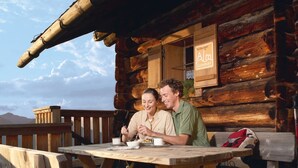 Eine Holzhütte mit Esstisch und Bänken, einem Schild mit der Aufschrift 'Salzburger Alm Sommer' und Blick auf die Berge im Hintergrund.