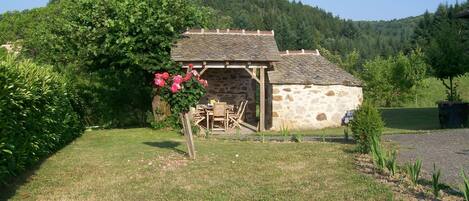 Ein Steinhaus mit Ziegeldach, draußen ein Holztisch und Stühle, umgeben von Grünpflanzen und einem Hügel im Hintergrund.