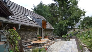 A stone house with a tiled roof, a patio with outdoor furniture, and a garden with various plants and a statue. - Cozy Apartment in Immerath near Fishing (Immerath)