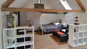A loft bedroom with a sloped ceiling, exposed wooden beams, a bed with a gray bedspread, a small desk with a lamp, and a white shelving unit. - Cozy Apartment in Immerath near Fishing (Immerath)