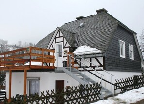 Ein Haus mit einem steilen Satteldach, einer Holztreppe, die zu einem Balkon führt, und einem umzäunten Garten.