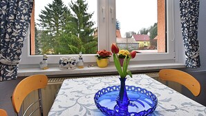 Ein Esstisch mit einer blauen Glasvase und Tulpen, ein Fenster mit Spitzenvorhängen und Blick auf Bäume und Gebäude.