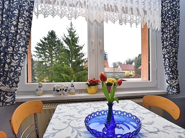 Uma mesa de jantar com um vaso de vidro azul e tulipas, uma janela com cortinas de renda e vista para árvores e edifícios lá fora.
