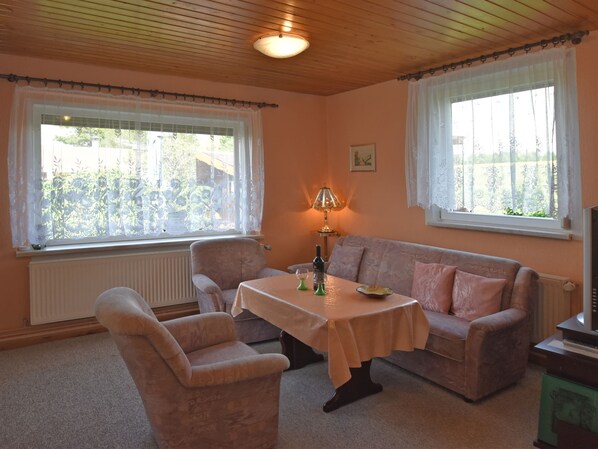 A cozy living room with a wooden ceiling, two windows with white curtains, a table with a lamp, a sofa, and a radiator.