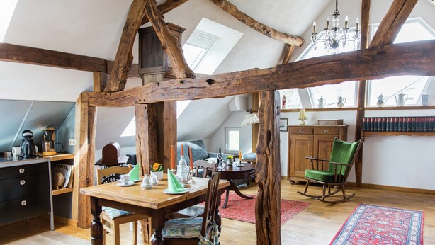 A loft-style room with exposed wooden beams, a dining table with chairs, a chandelier, and a red patterned rug. - Apartment in Schleiden (Schleiden)