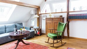 A cozy loft-style room with a high ceiling, exposed wooden beams, a blue sofa, a rocking chair, a wooden cabinet, and a bookshelf filled with books. - Apartment in Schleiden (Schleiden)