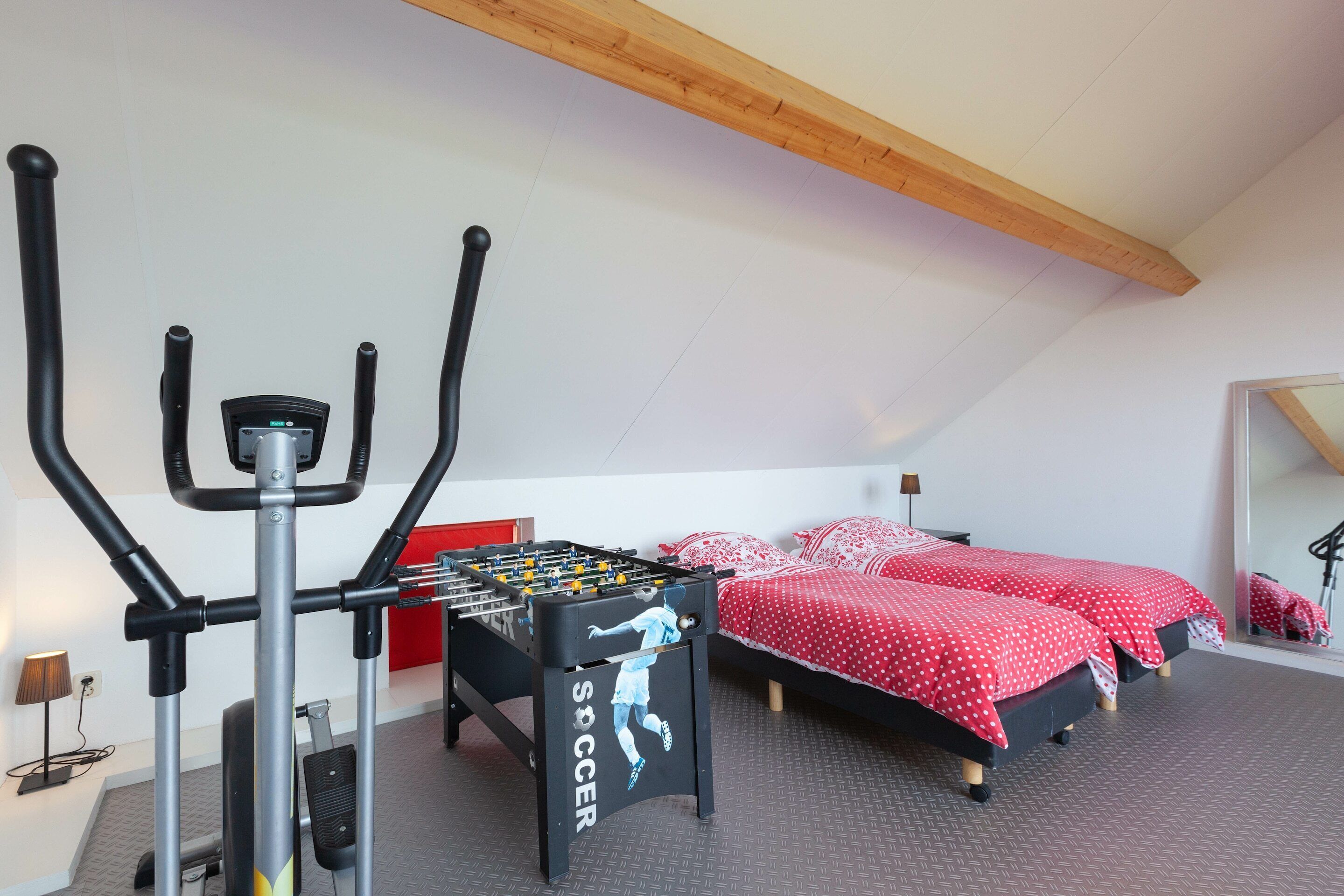 Una habitación con mesa de futbolín, equipo de ejercicio, dos camas con ropa de cama a lunares rojos y un espejo grande.