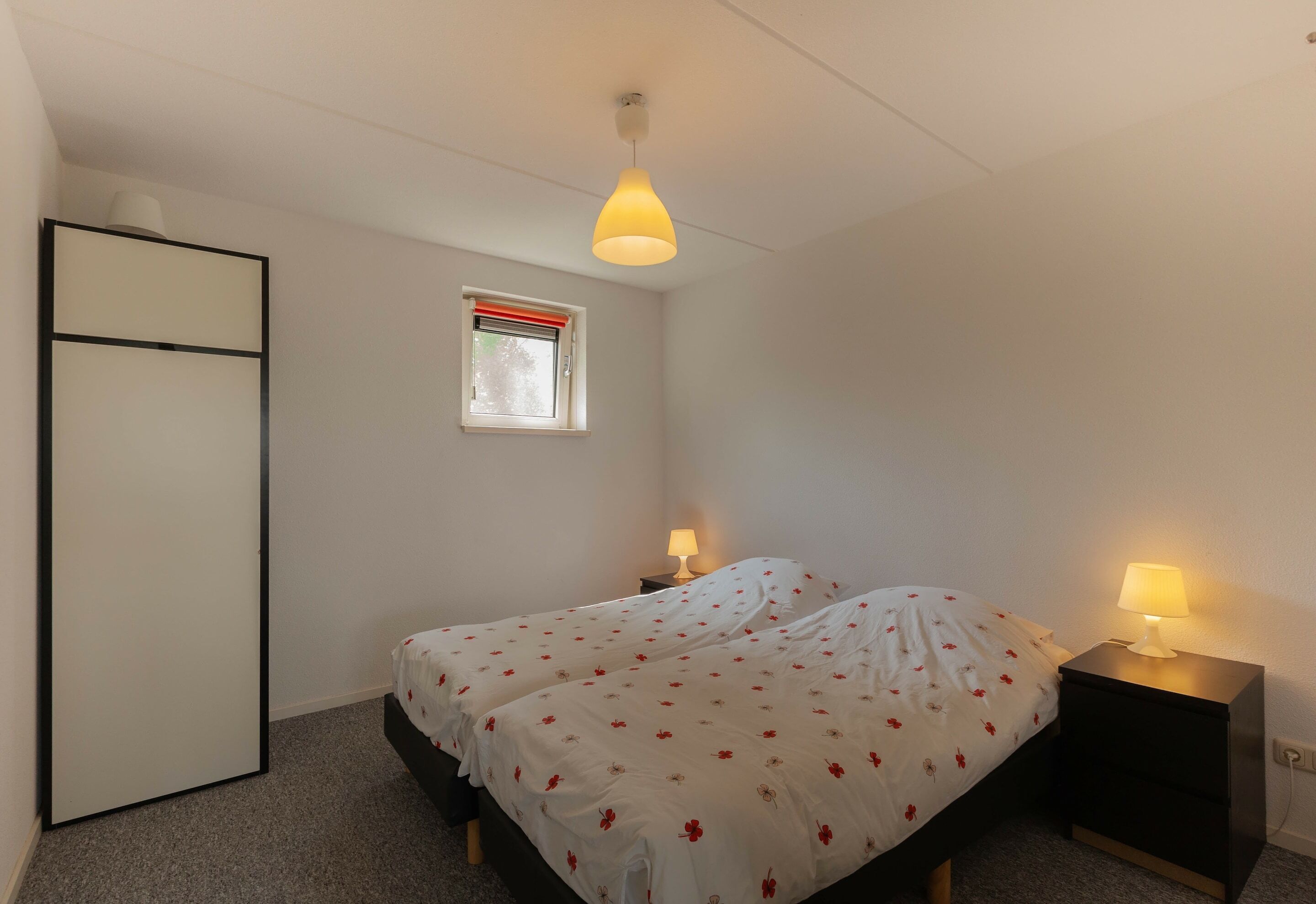A bedroom with a bed, two bedside tables with lamps, a window with a red blind, and a door.