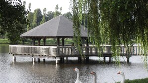 Ein Pavillon über einem See, mit drei Gänsen am Ufer.