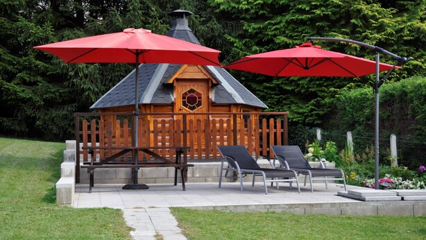 Ein hölzerner Pavillon mit Schornstein, zwei roten Sonnenschirmen und zwei Liegestühlen.