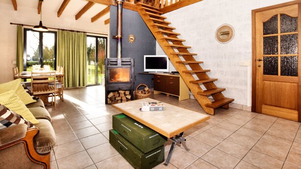 A cozy living room with a wooden staircase, a fireplace, a dining area, and a large window. - Holiday Home in Peaceful Setting and Large Garden (Rendeux)