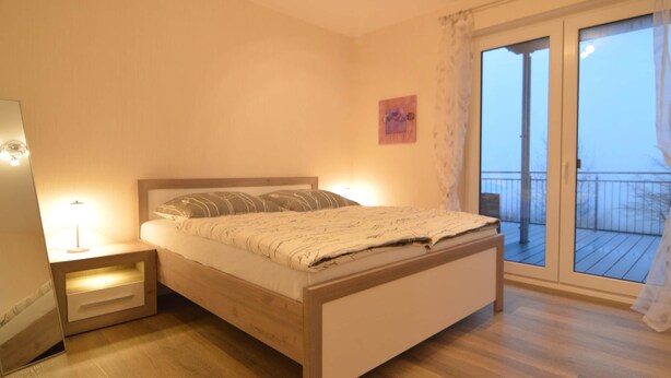 Una camera da letto con un letto di legno, un comodino con una lampada, un'ampia finestra vista balcone e un piccolo quadro appeso al muro.