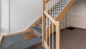 Uma escadaria com corrimãos de madeira e degraus de metal, conduzindo a um patamar com uma grade metálica e um tapete de madeira.