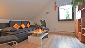 A modern living room with a dark grey sofa, orange cushions, a white coffee table with a vase of flowers, and a large window with a view of greenery. - Gemutliches Familienapartment (Werda)