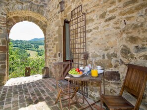 A rustic stone room with a table set for breakfast, a view of a vineyard, and a wooden bench.