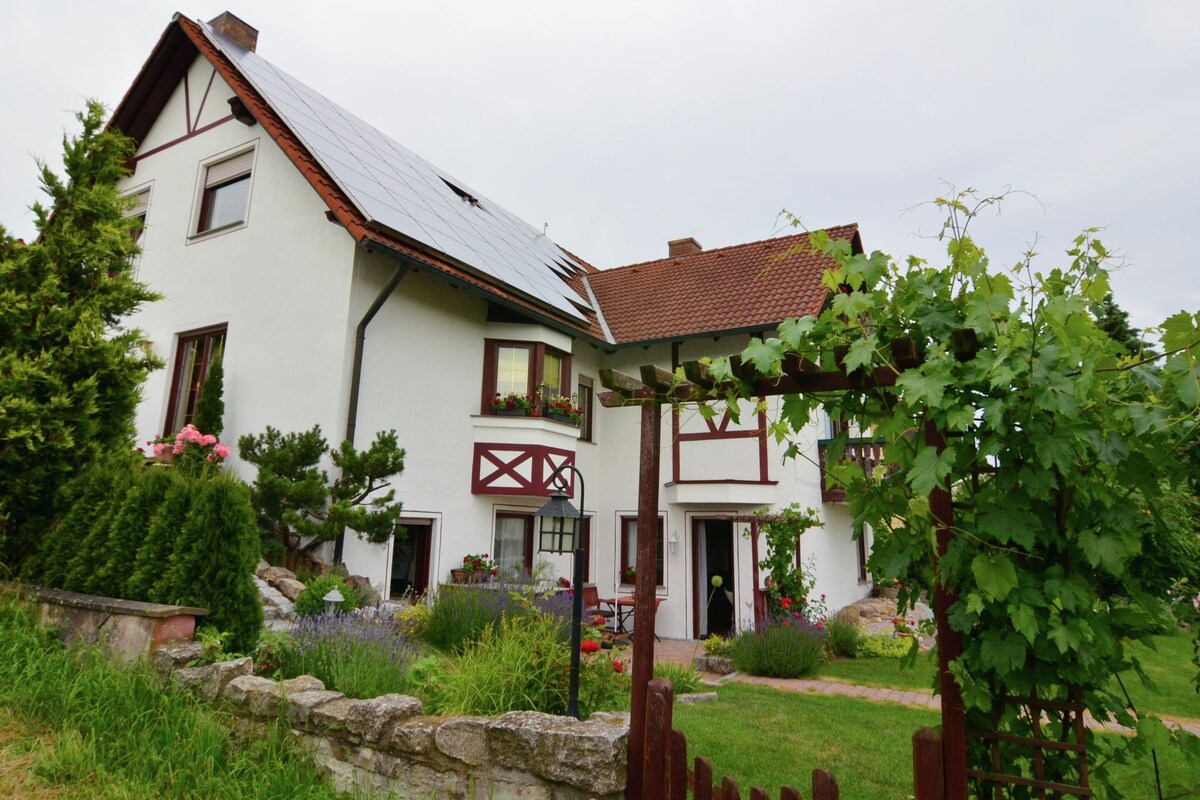 A two-story house with a sloped roof, a stone wall, and a garden with various plants and flowers.