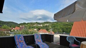 Una terraza en la azotea con sillones y una mesa, con vistas a un pueblo de casas con tejados rojos y colinas verdes.