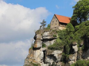 A historic building perched atop a rocky cliff with a red roof, surrounded by lush greenery.