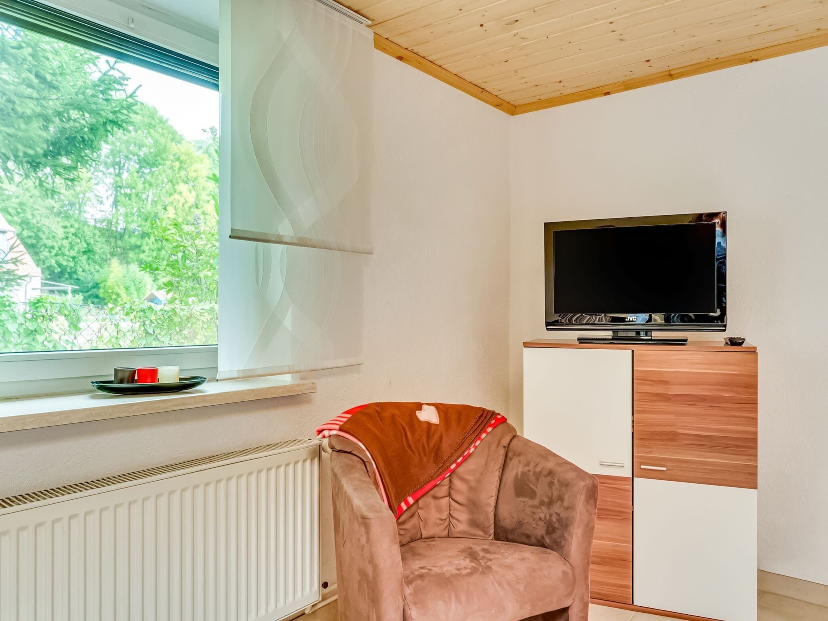 A room with a radiator, a window with blinds, a television on a wooden cabinet, and a chair with a blanket.