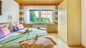 A bedroom with a wooden ceiling, a window with curtains, a bed with multiple pillows, and a wardrobe.