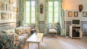 A room with a floral-patterned sofa, a wooden table, a vintage clock, a television on a stand, and framed pictures on the wall. - Villa in Grimaud Near Tennis Court & Pool (Montbrun-les-Bains)