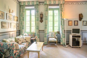 A room with a floral-patterned sofa, a wooden table, a vintage clock, a television on a stand, and framed pictures on the wall. - Villa in Grimaud Near Tennis Court & Pool (Montbrun-les-Bains)