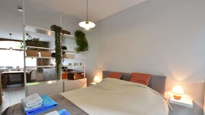 A modern bedroom with a large bed, bedside lamps, a desk, and a shelving unit with plants. - Apartment in Ieper With Terrace (Ypres)