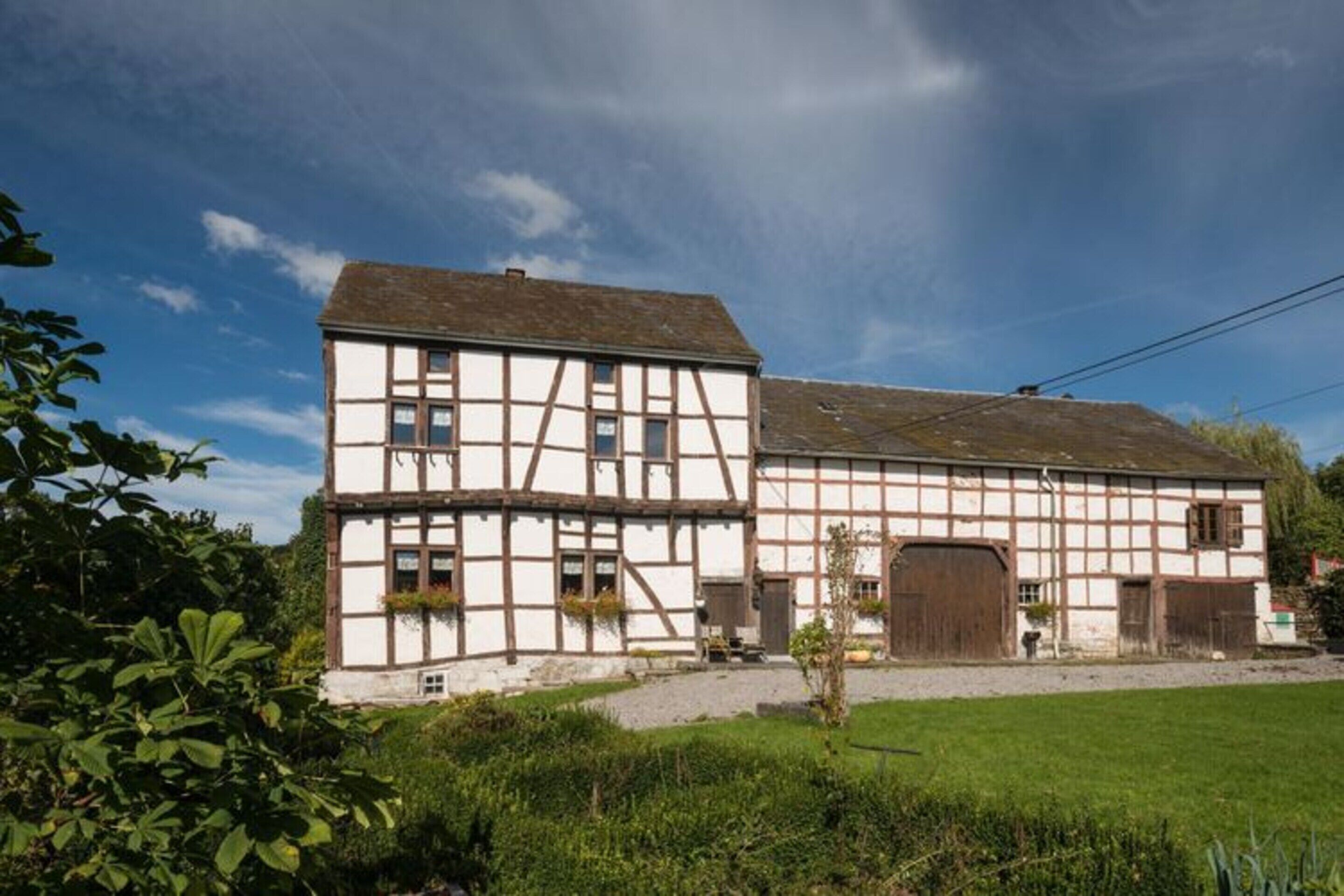 Une maison traditionnelle à colombages, dotée d’une grande porte en bois et de fenêtres, entourée de verdure.