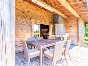 A wooden cabin with a dining area featuring a table and wicker chairs, a stone fireplace, and a wooden deck. - Traditional Chalet with Garden near Malmedy & Hautes-Fagnes (Malmedy)