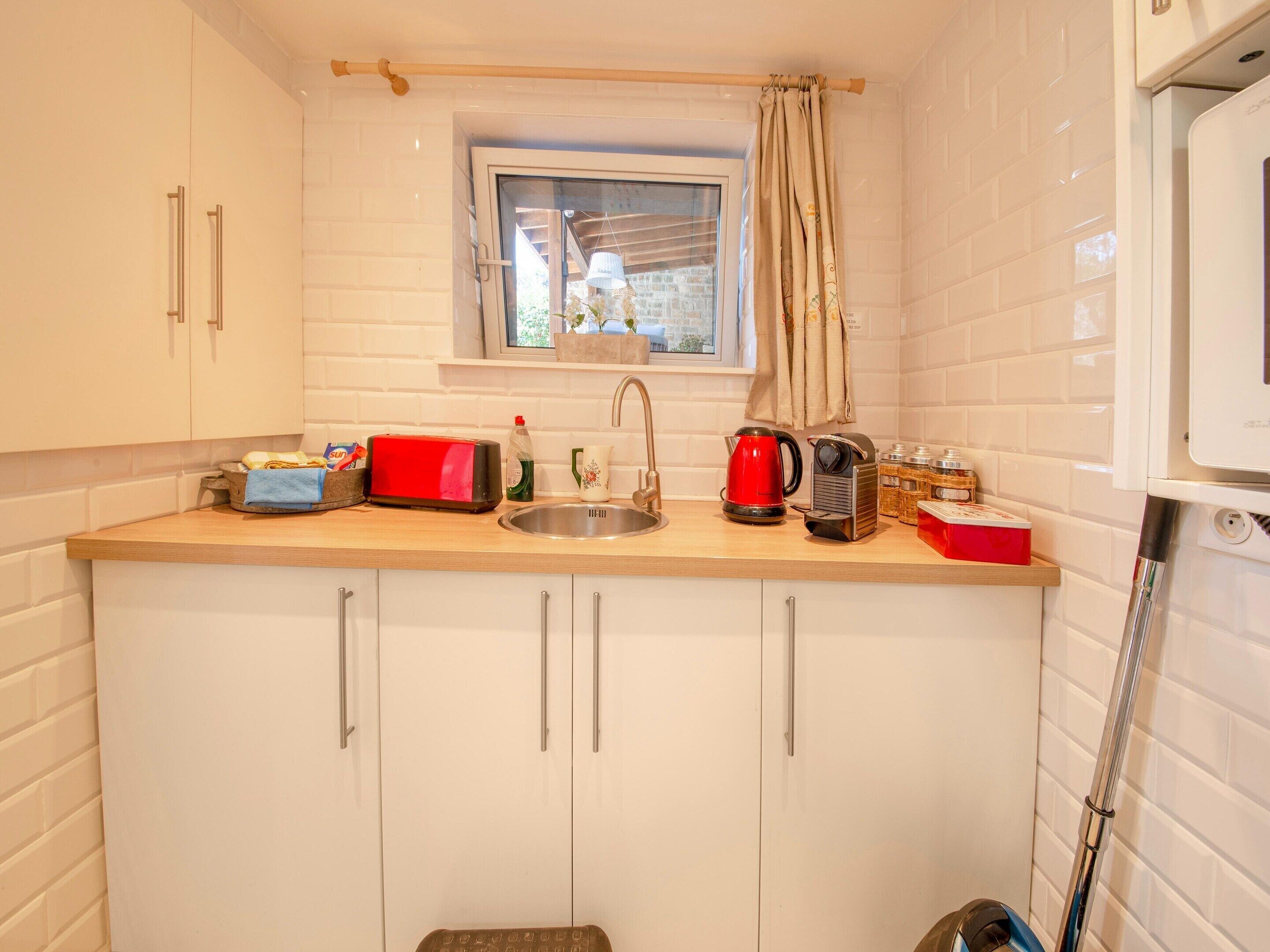 Een kleine keuken met witte kastjes, een houten aanrecht, een raam met uitzicht, een spoelbak, een rode waterkoker en een broodrooster.
