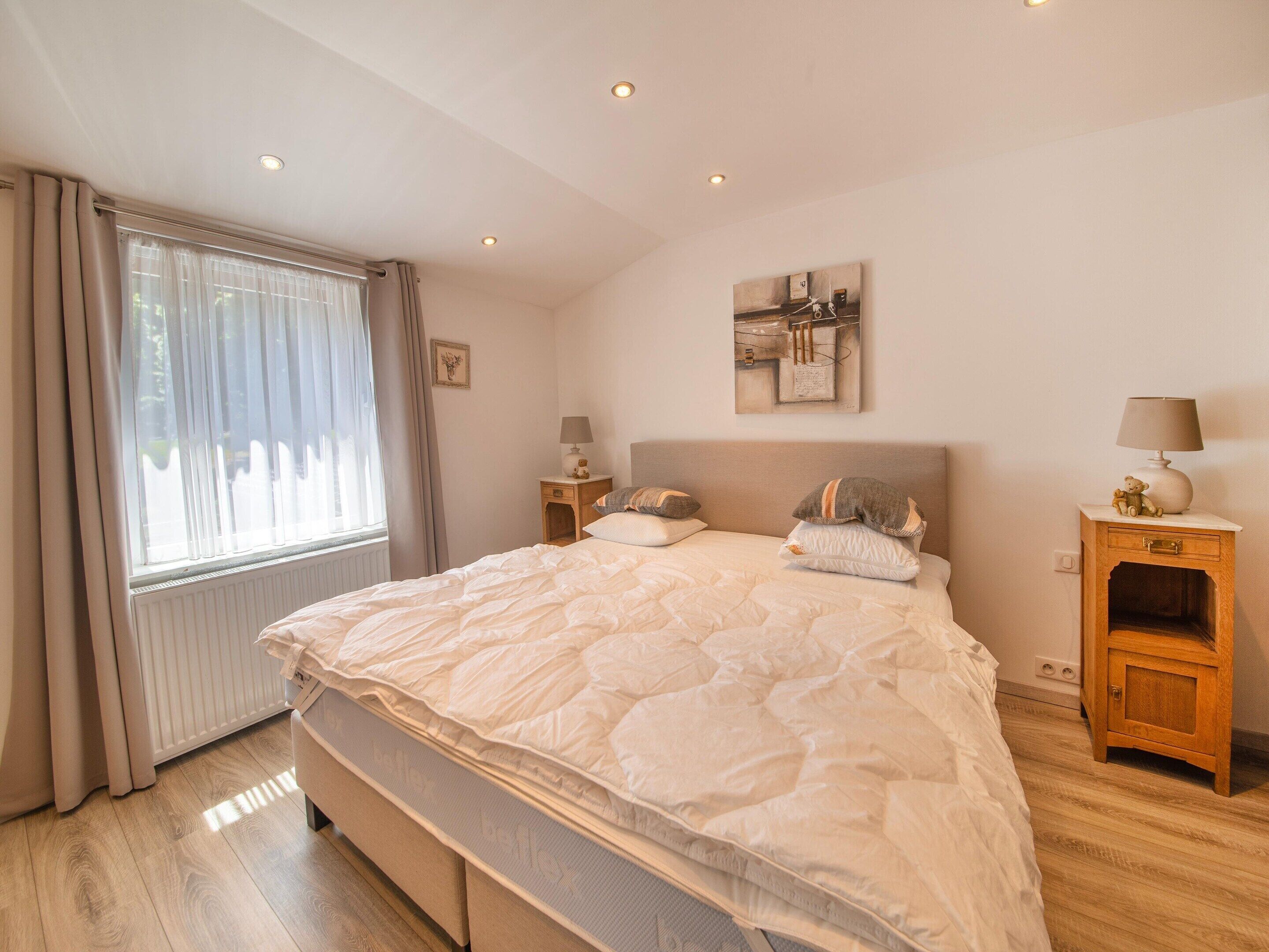 A bedroom with a large bed, two bedside tables, a window with curtains, a wall-mounted artwork, and a radiator.