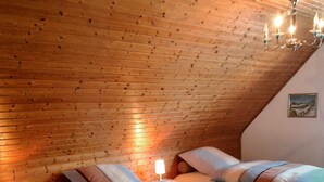 Tunnelmallinen makuuhuone, jossa on kaksi sänkyä, puupalkkinen katto, yöpöytä lampun kanssa ja seinällä oleva kehystetty kuva.