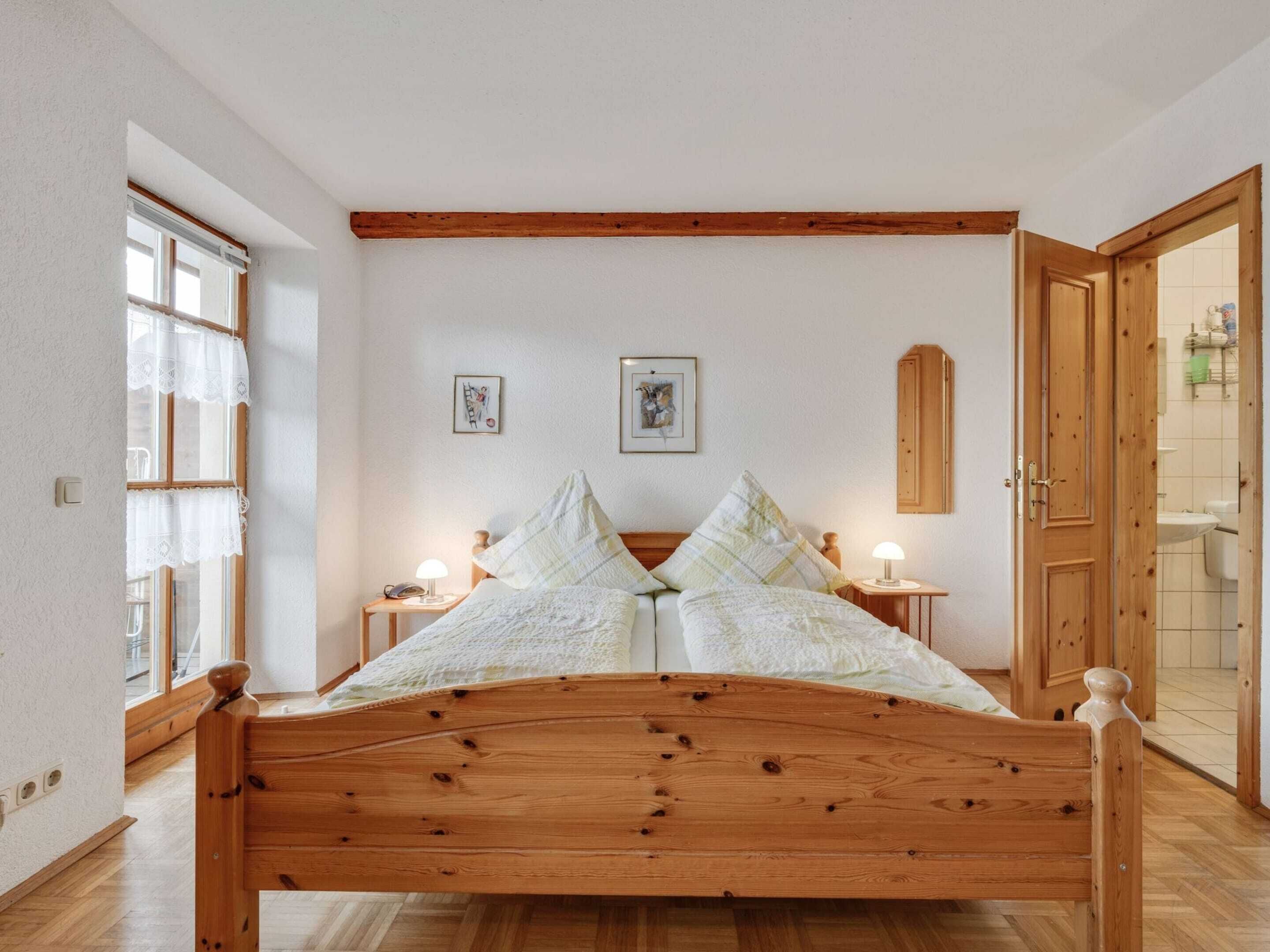 A bedroom with a wooden bed, two bedside tables with lamps, a window with lace curtains, and a door leading to a bathroom.