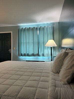 A bedroom with a bed, a window with curtains, a lamp, and a door.