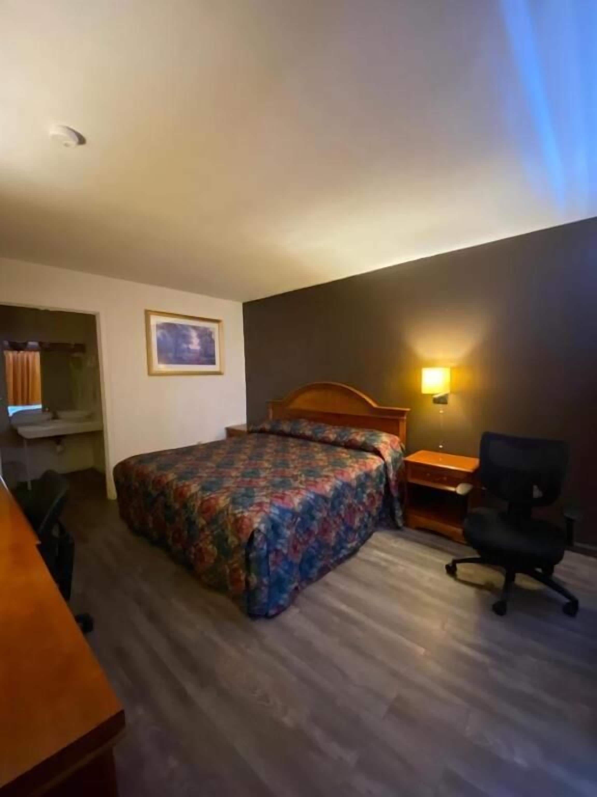 Ein Hotelzimmer mit Bett, Schreibtisch, Stuhl, Nachttisch, Lampe und einem Bild an der Wand.