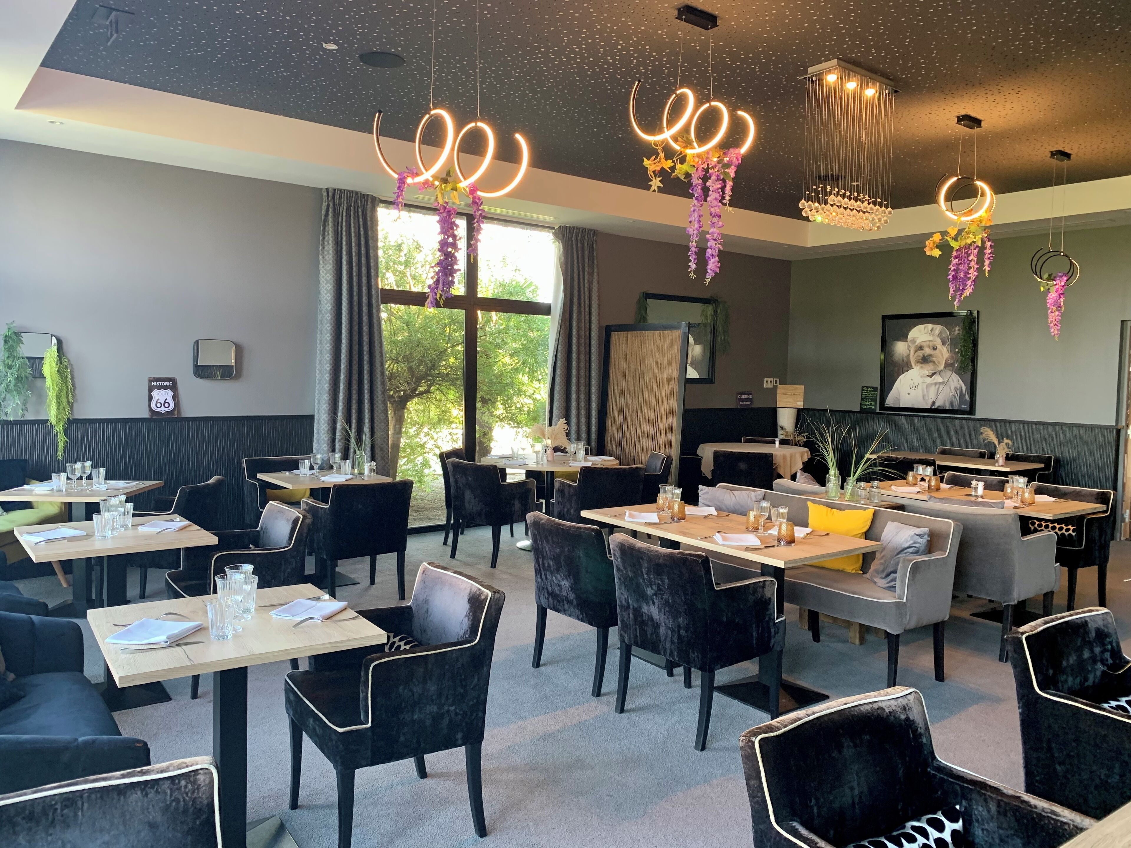 A restaurant with multiple tables and chairs, a large window with a view of trees, and decorative hanging lights.